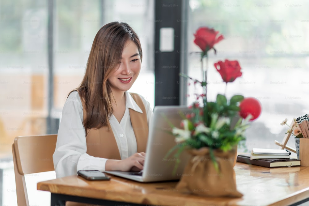 Belle jeune femme asiatique assise dans un café à l’aide d’un ordinateur portable. Heureuse jeune femme d’affaires assise à la table dans un café avec un ordinateur à onglet.