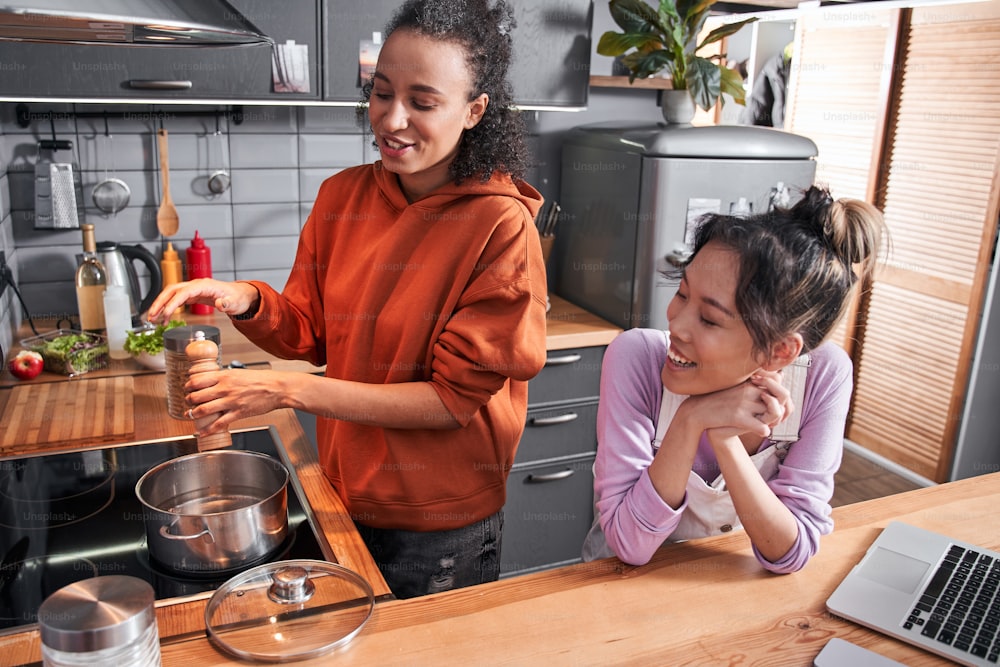 Girls chatting with pleasure during the cooking at home. High quality photoPortrait view of the multiracial woman salt pasta and sprinkle with species while preparing dinner with her best friend at the kitchen. Girls chatting with pleasure during the cooking. Coolinary concept. Stock photo