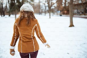 ニット帽とシープスキンのコートを着たミトンを着た中年女性が、冬の都市公園を歩いている後ろから見た。