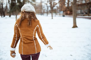 Seen from behind middle aged woman with mittens in a knitted hat and sheepskin coat walking outside in the city park in winter.