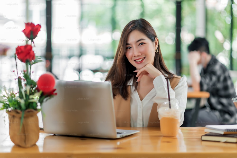 Belle jeune femme asiatique assise dans un café à l’aide d’un ordinateur portable. Heureuse jeune femme d’affaires assise à la table dans un café avec un ordinateur à onglet.