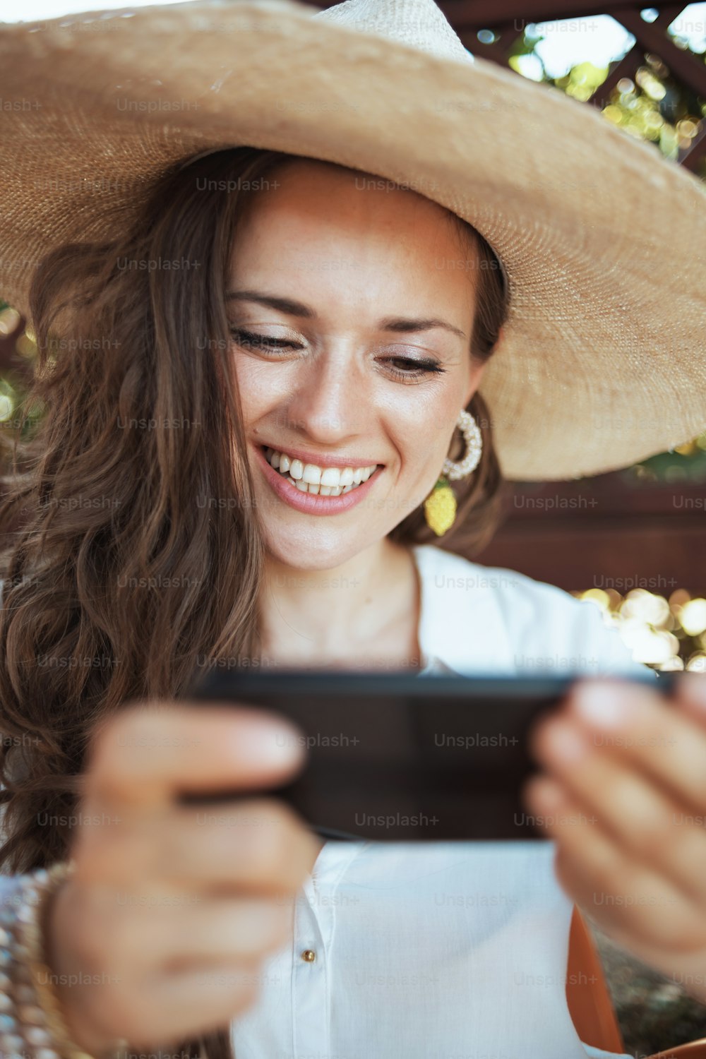Lächelnde moderne 40-jährige Hausfrau im weißen Hemd mit Hut per Smartphone-App auf der Terrasse des Guest House Hotels.
