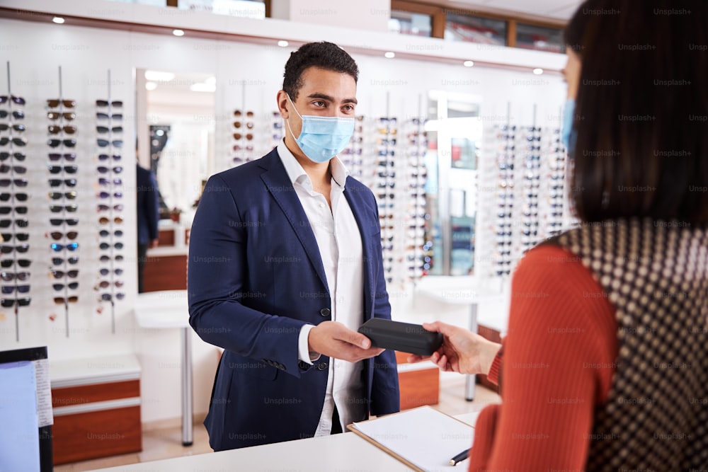 Young male customer in a face mask taking a spectacle case from the optician hands