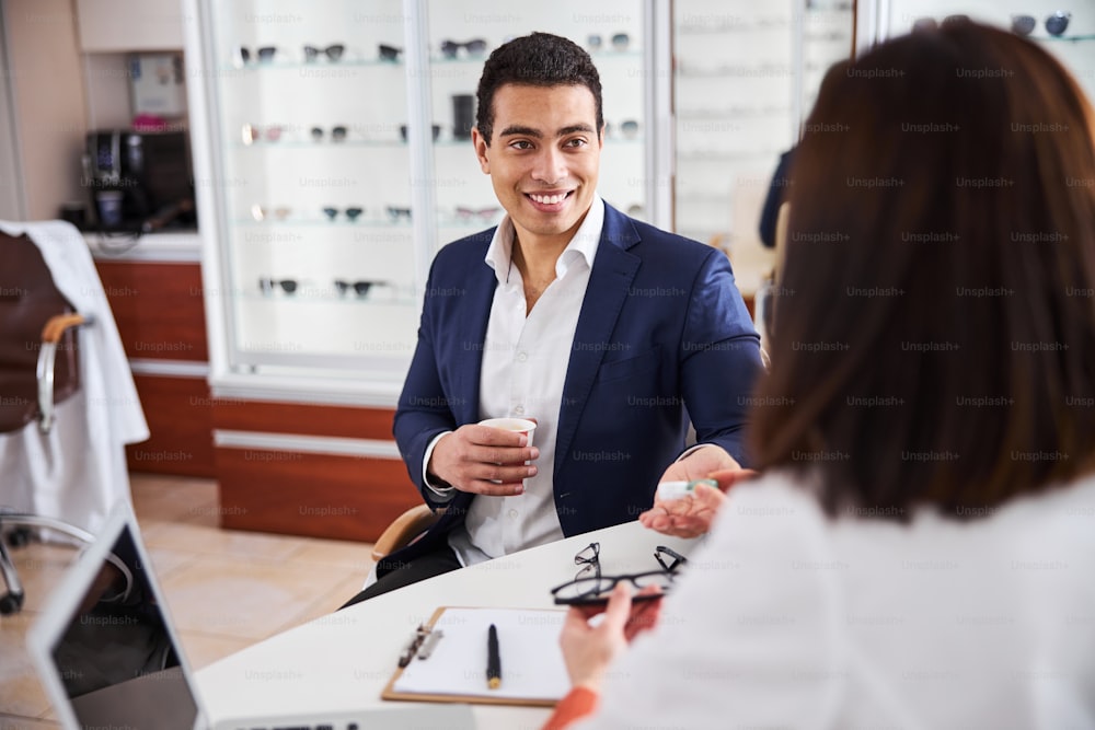 Attractive smiling young male customer looking at a dark-haired female eyewear consultant at the desk