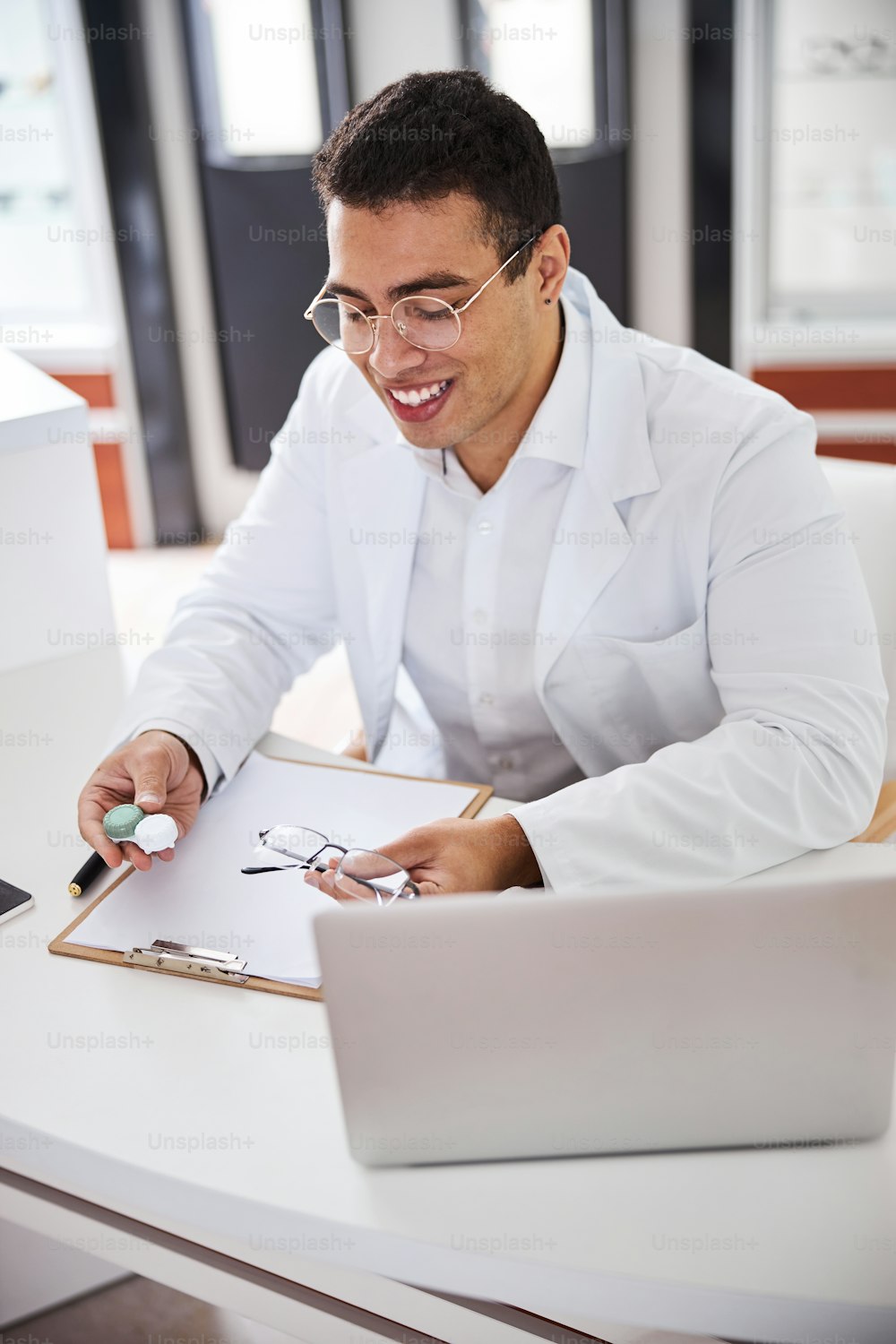 Contented bespectacled optician with a pair of glasses and the contacts sitting at the desk