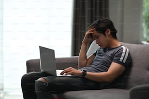 Young Asian man sitting on couch with laptop and feeling tired from working online at home.
