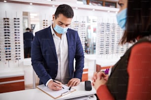 Focused customer in a face mask filling out a blank in the presence of an optician