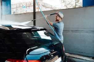 Giovane con cappello che lava la sua auto durante il giorno alla stazione di autolavaggio usando acqua ad alta pressione.