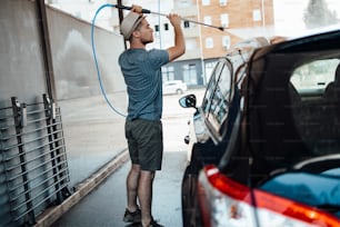 Jovem com chapéu lavando seu carro durante o dia na estação de lavagem de carros usando água de alta pressão.