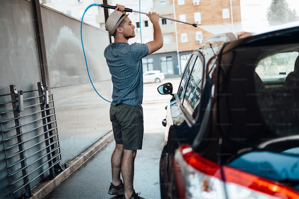 Jovem com chapéu lavando seu carro durante o dia na estação de lavagem de carros usando água de alta pressão.