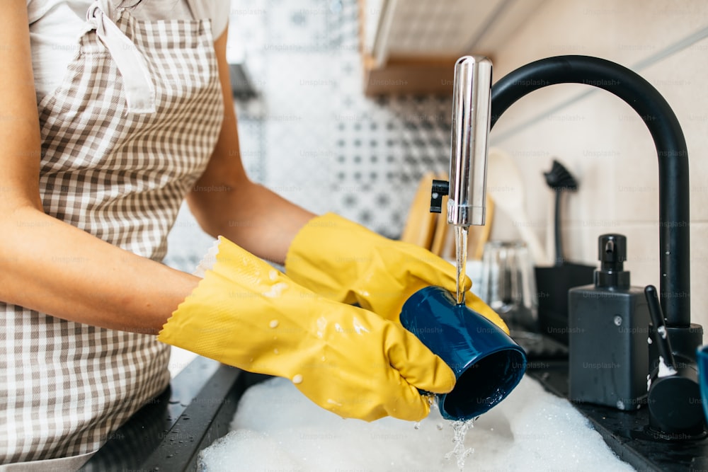 Mulher adulta jovem com luvas de proteção amarelas lavando seus pratos na pia da cozinha. Rotina de higiene doméstica e doméstica.