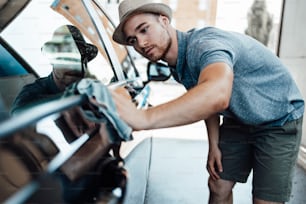 Giovane bell'uomo con cappello che pulisce l'auto con lo straccio, concetto di dettaglio dell'auto (o valeting).