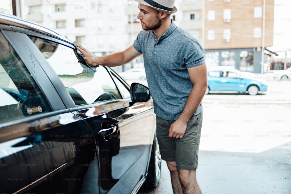 Joven guapo con sombrero limpiando el coche con el trapo, concepto de detalle del coche (o valeting).