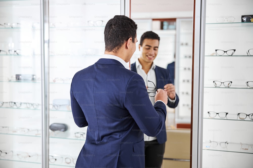 Back view of a smiling buyer with a pair of glasses standing before the mirror