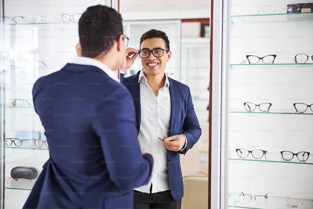 Merry customer in trendy spectacles standing in front of the mirror in an optical shop