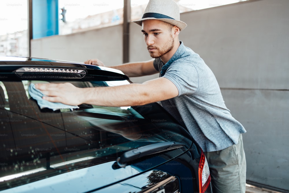 ぼろきれの車を掃除する帽子をかぶった若いハンサムな男、車のディテール(またはバレー)のコンセプト。
