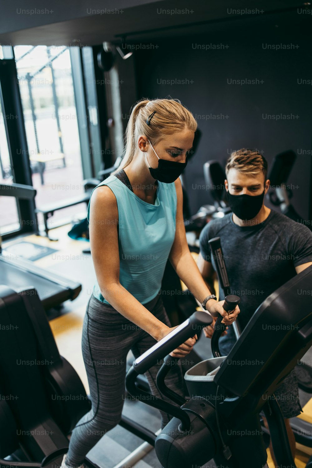 Young fit and attractive woman at body workout in modern gym together with her personal fitness instructor or coach. They keeping distance and wearing protective face masks. Coronavirus world pandemic and sport theme.