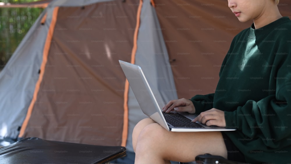 Young Asian woman using laptop near tent during camping in field.