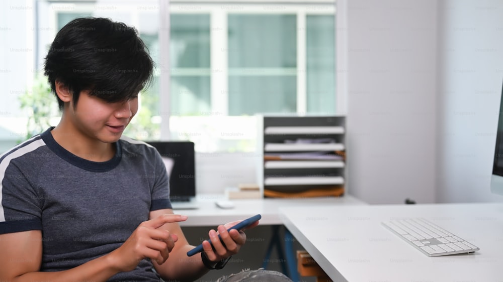 Young Asian man freelance sitting at his modern home office and using smart phone.