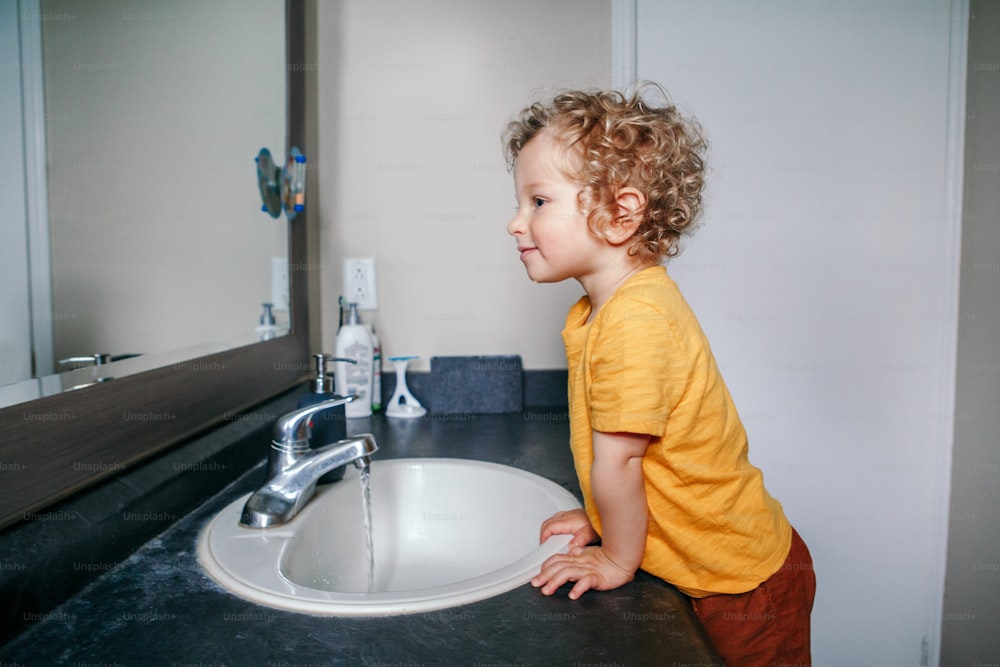 Little Caucasian boy toddler washing hands in bathroom at home. Health hygiene and morning routine for children. Cute funny child playing with water. Kid looking in mirrow in washroom.