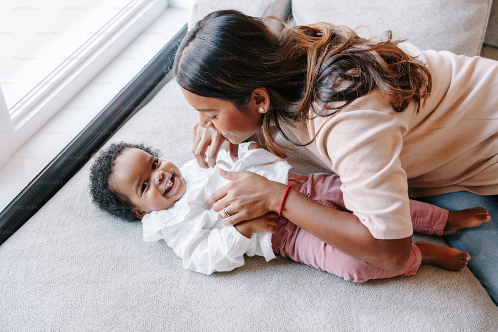 黒の女の赤ちゃんの娘と遊ぶ幸せな笑顔の若いインドの母。家族、混血、お母さん、子供が家で一緒に抱きしめている。幼児の子供との本物の率直なライフスタイル。