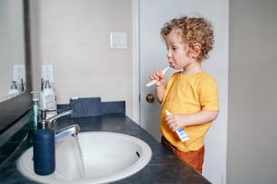 Little Caucasian boy toddler brushing teeth in bathroom at home. Health hygiene and morning routine for children. Cute funny child washing in toilet. Kid looking in mirror in washroom.