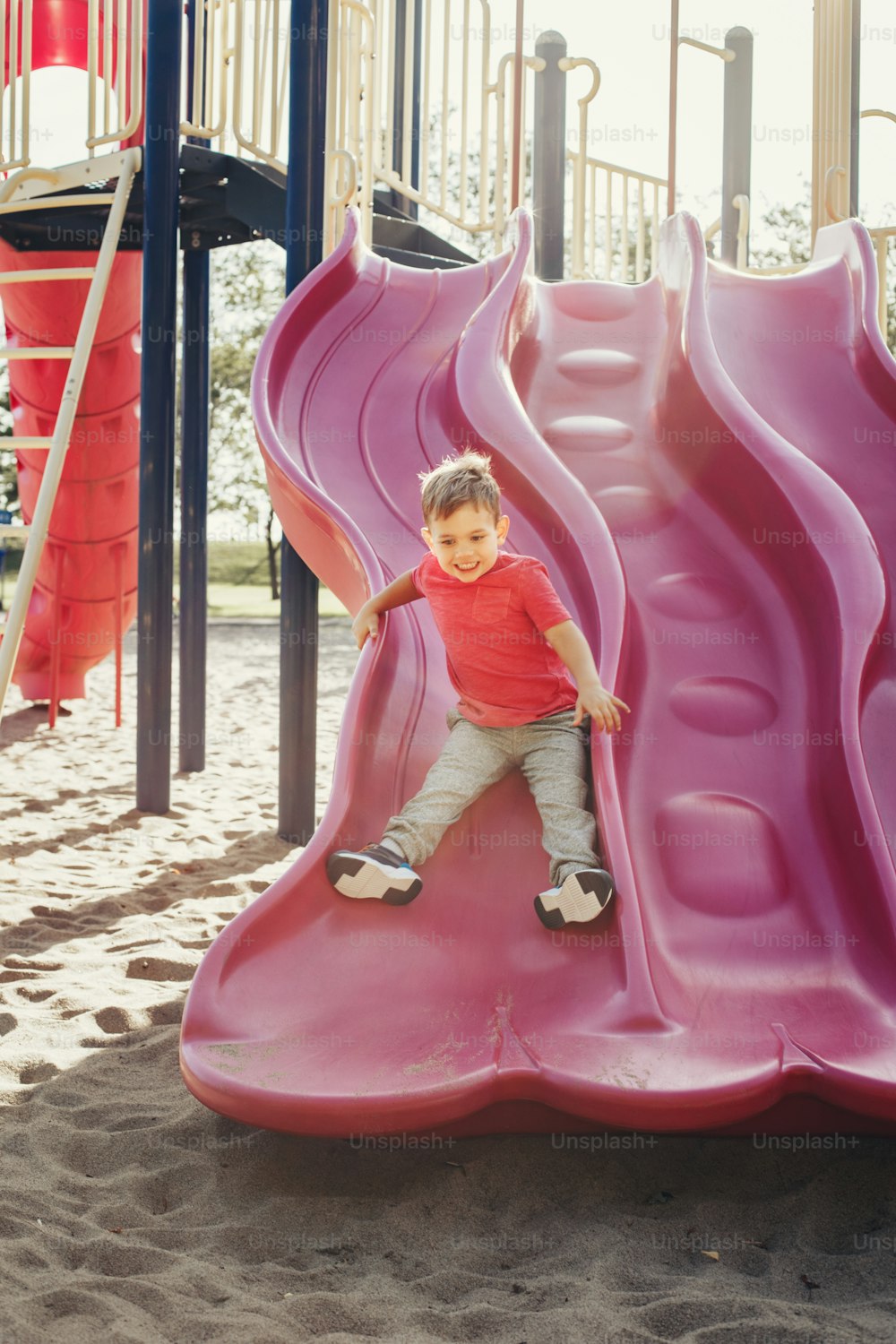 Ragazzo caucasico sorridente felice attivo che scivola sul parco giochi. Bambino nel cortile della scuola all'aperto in una giornata di sole estiva. Bambino che si diverte all'aperto sul terreno di gioco. Attività sportiva stagionale per bambini.