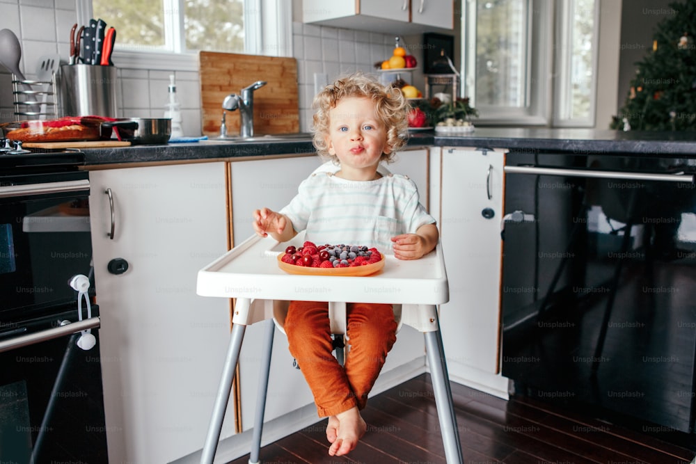 Cute Caucasian baby boy eating ripe red fruits at home. Funny child kid sitting high chair with fresh berries in kitchen. Supplementary healthy finger food for toddler kids.