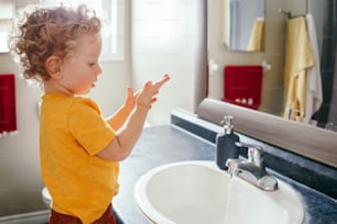 Little Caucasian boy toddler washing hands in bathroom at home. Health hygiene and morning routine for children. Cute funny child playing with water. Kid learning exploring his body.