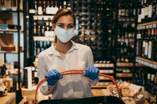 Beautiful young and elegant woman with face protective mask and gloves buying healthy food and drink in a modern supermarket or grocery store. Pandemic or epidemic lifestyle and consumerism concept.