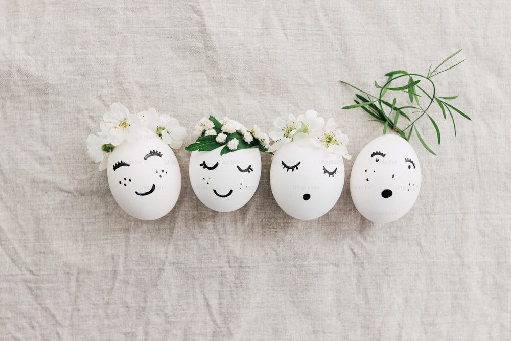 Natural eggs with drawn funny faces in cute floral crowns top view on linen fabric with blooming spring petals and green leaves. Happy Easter! Eco friendly holiday concept. Space for text