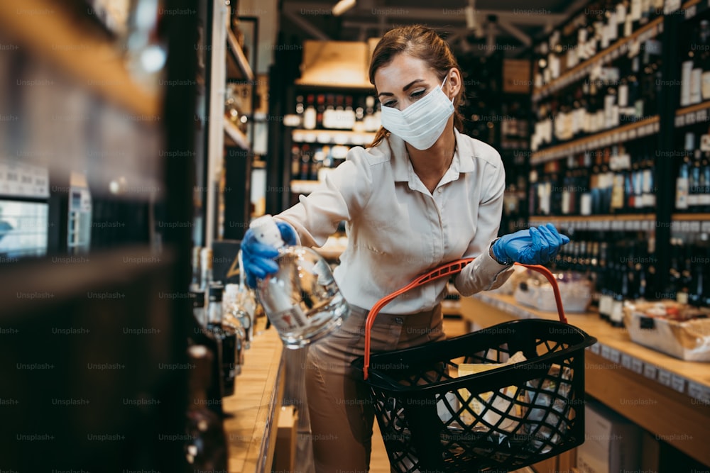 Beautiful young and elegant woman with face protective mask and gloves buying healthy food and drink in a modern supermarket or grocery store. Pandemic or epidemic lifestyle and consumerism concept.