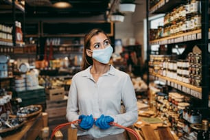 Beautiful young and elegant woman with face protective mask and gloves buying healthy food and drink in a modern supermarket or grocery store. Pandemic or epidemic lifestyle and consumerism concept.