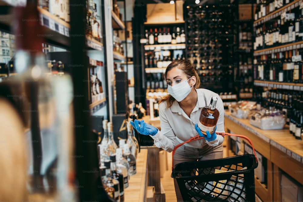 Beautiful young and elegant woman with face protective mask and gloves buying healthy food and drink in a modern supermarket or grocery store. Pandemic or epidemic lifestyle and consumerism concept.