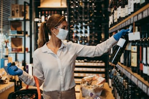 Beautiful young and elegant woman with face protective mask and gloves buying healthy food and drink in a modern supermarket or grocery store. Pandemic or epidemic lifestyle and consumerism concept.
