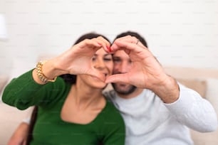 Beautiful young couple at home is making heart sign with hands, smiling and looking at camera. Celebrating Saint Valentine's Day.