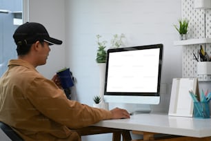 그의 홈 오피스에서 현대 컴퓨터에서 온라인으로 작업하는 젊은 아시아 남자의 측면 모습.