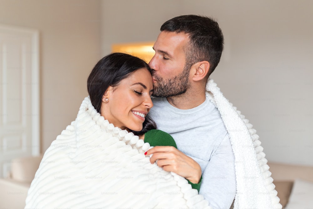Pareja romántica de vacaciones de invierno. Hombre y mujer de pie juntos en una habitación de hotel envueltos en una manta. Pareja abrazándose y sonriendo.