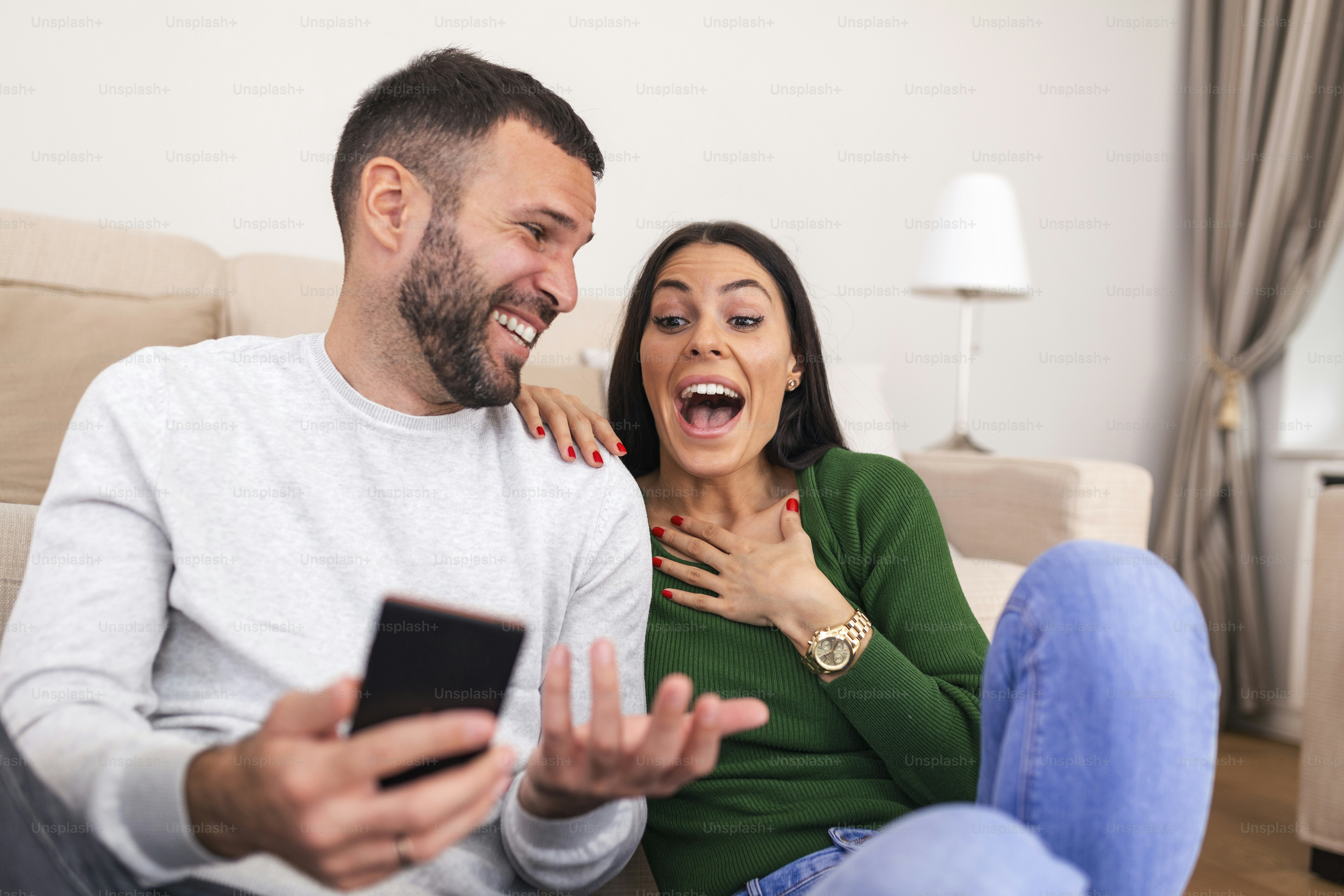 Smiling young couple sit relax on couch in living room have fun using smartphone together, happy Caucasian man and woman rest on sofa watch video on cellphone, make self-portrait picture on cell