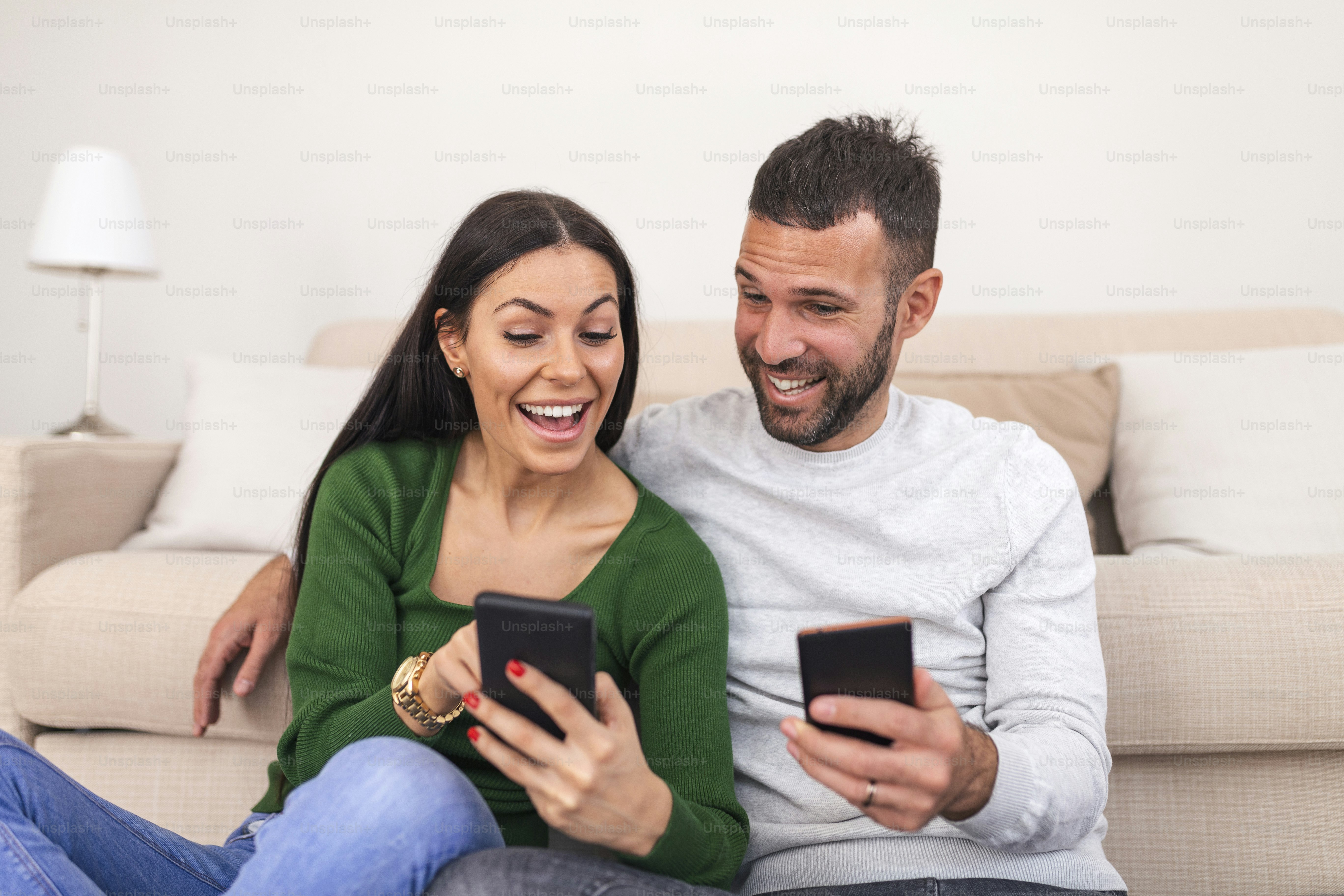 Beautiful young couple in love at home, sitting at the living room, pointing at mobile phone. young couple surfing social media together and laughing at funny posts