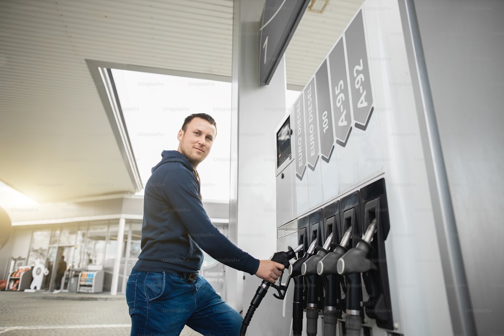 Young handsome man in casual clothes, refueling his car with gasoline at a gas station, taking filling gun from petrol panel.