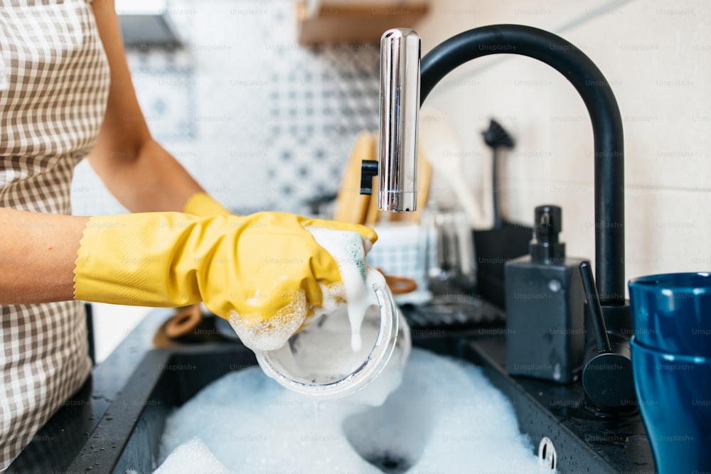 Jeune femme adulte avec des gants de protection jaunes lavant sa vaisselle sur l’évier de la cuisine. Routine d’hygiène domestique et domestique.