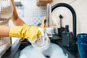 Giovane donna adulta con guanti protettivi gialli che lava i piatti sul lavello della cucina. Routine di igiene domestica e domestica.