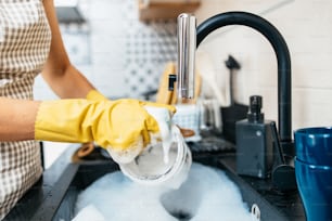 Young adult woman with yellow protective gloves washing her dishes on kitchen sink. Household and home hygiene routine.