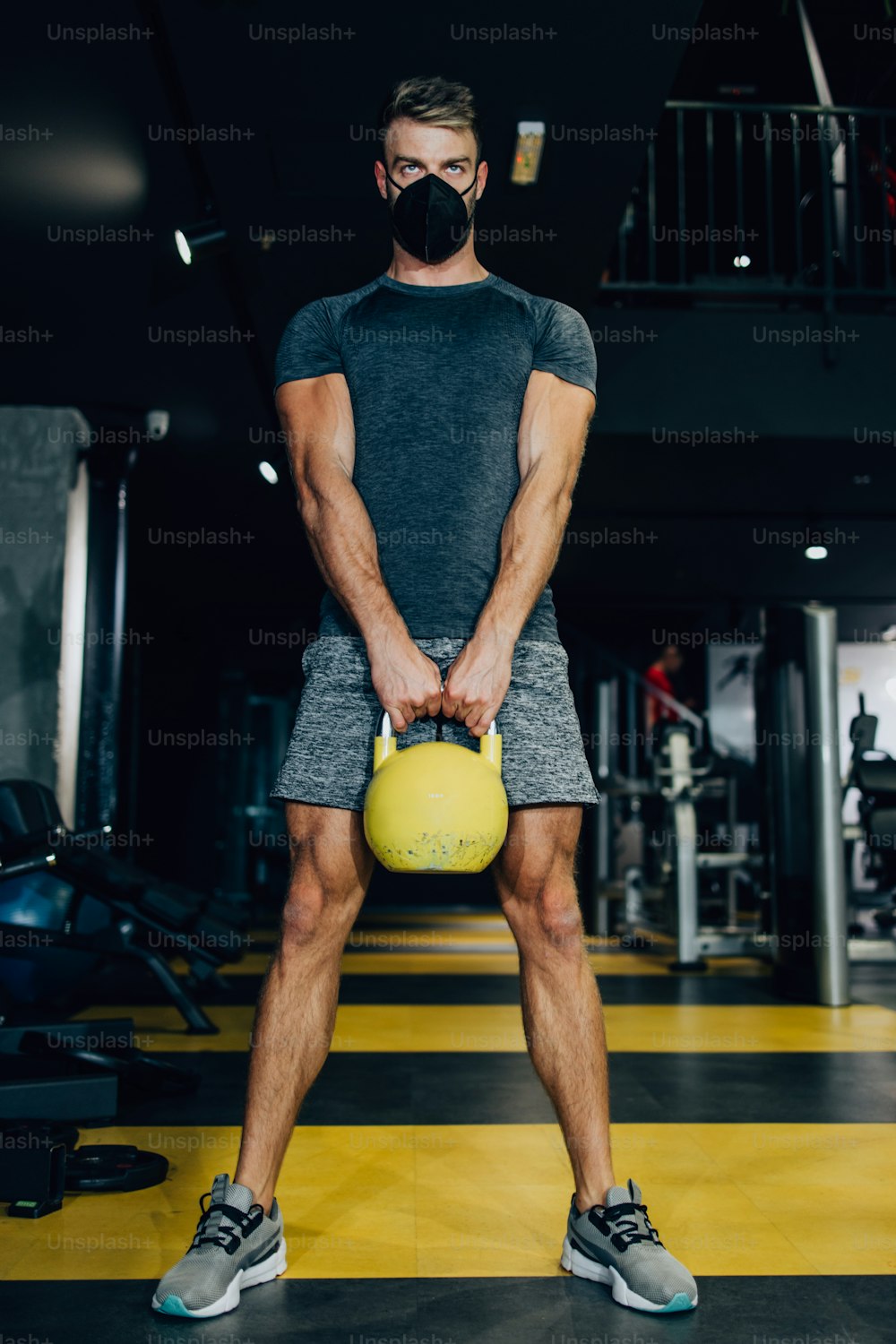 Good looking handsome male athlete with protective face mask exercising in modern fitness gym. Dark muddy light with strong shadows. Pandemic sports indoors concept.