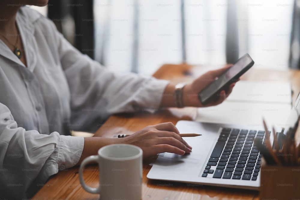 Cropped shot of businesswoman working with laptop computer and  searching information smart phone at office desk.