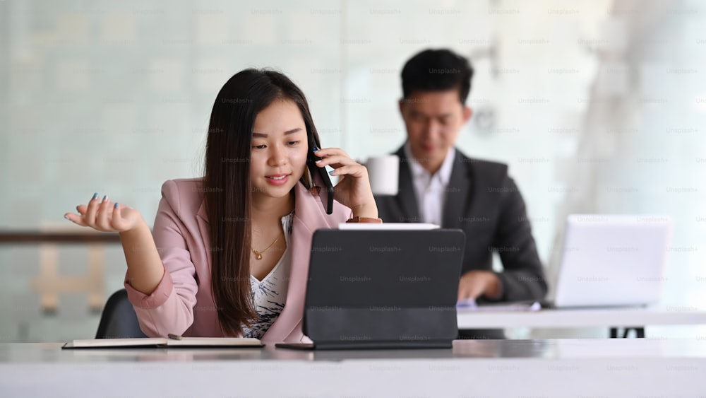 Junge asiatische Geschäftsfrau, die mit Tablet-Computern arbeitet und im Büro ein Geschäftsgespräch auf dem Handy führt.