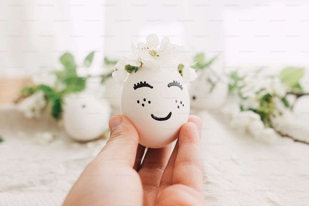 Happy Easter and Easter hunt concept ! Hand holding easter egg with drawn sleeping face in floral wreath on background of natural eggs and petals on linen fabric in soft light. Space for text