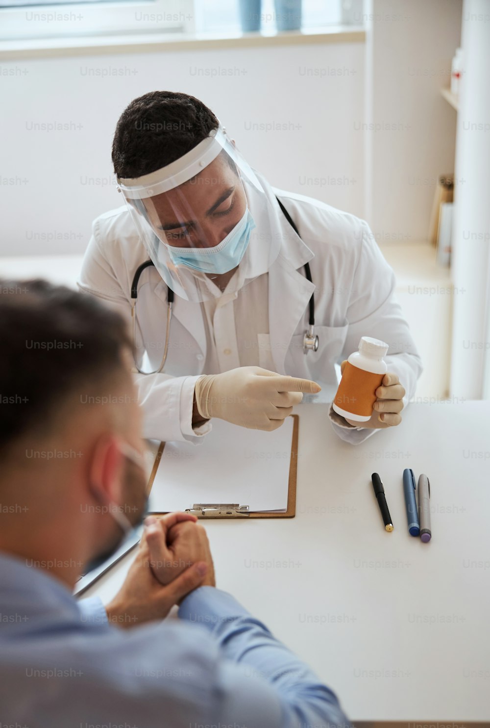 Retrato de cintura para arriba del médico masculino seguro de sí mismo en máscara protectora y uniforme blanco discutiendo con el paciente sobre la prescripción en el consultorio médico