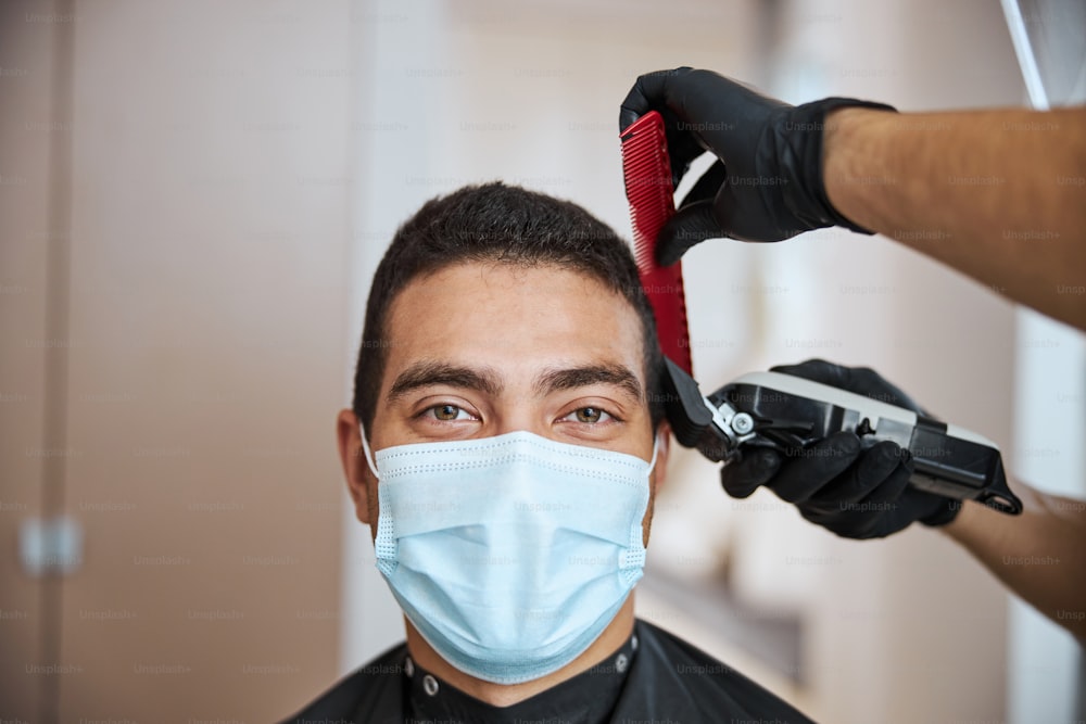 Close up portrait of handsome Indian man in protective mask sitting in male salon while looking at the photo camera while hairdresser cutting his hair